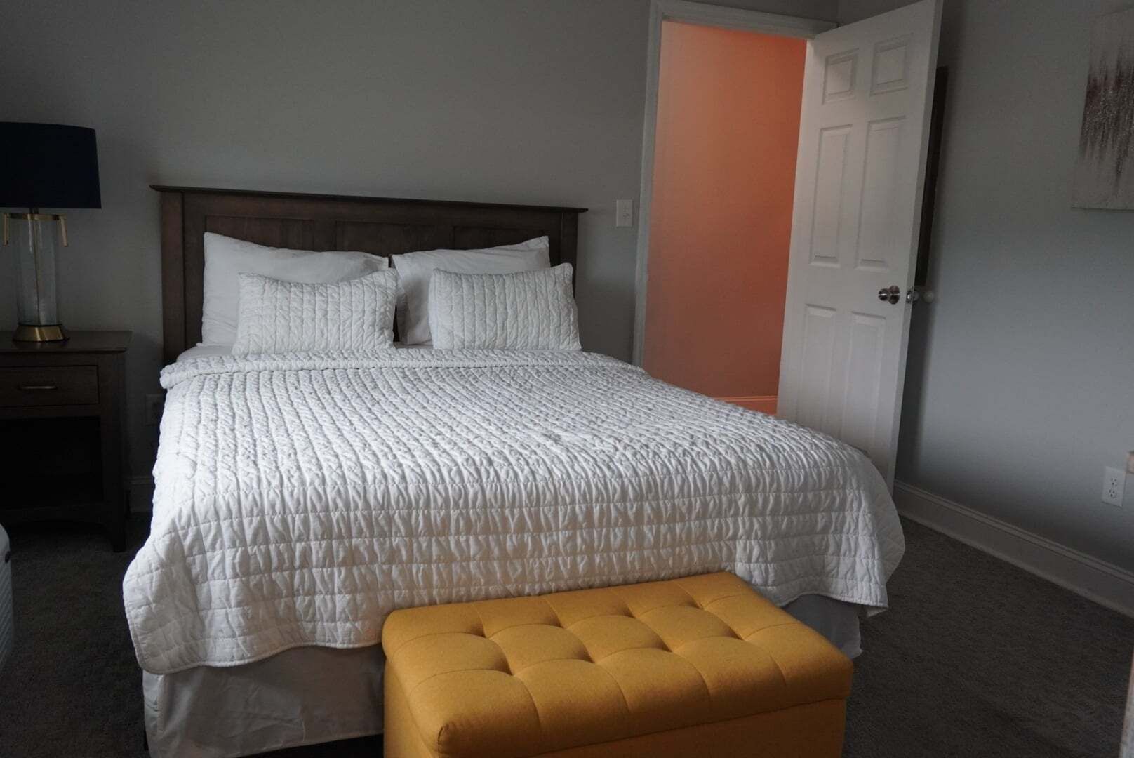 A bedroom with a bed, a nightstand, a lamp, a door, and a bench.