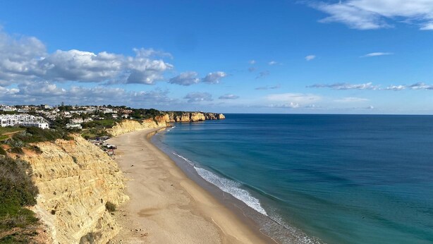Ein Küstenstrand mit sandigem Ufer, eine Klippe mit Gebäuden und einem strahlend blauen Himmel.