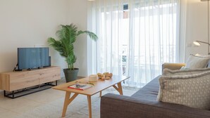 Ein modernes Wohnzimmer mit einer Couch, einem Couchtisch, einem Fernseher, einer Topfpflanze und transparenten Vorhängen.