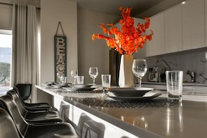 A modern kitchen with a dining area, featuring a central island with a vase of orange flowers, a 'BEACH' sign, and a view of the outdoors. - Prime Aruba Location: 2BR Condo near Palm Beach (Noord)