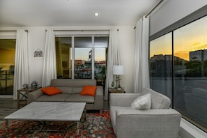 A modern living room with a sofa, armchair, and a coffee table. There is a large window with a city view. - Stylish 2BR Condo w/ Pool near Palm Beach (Noord)