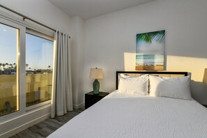 A hotel room with a large window, a bed with white bedding, a nightstand with a lamp, and a wall-mounted painting of a beach scene. - Stylish 2BR Condo w/ Pool near Palm Beach (Noord)