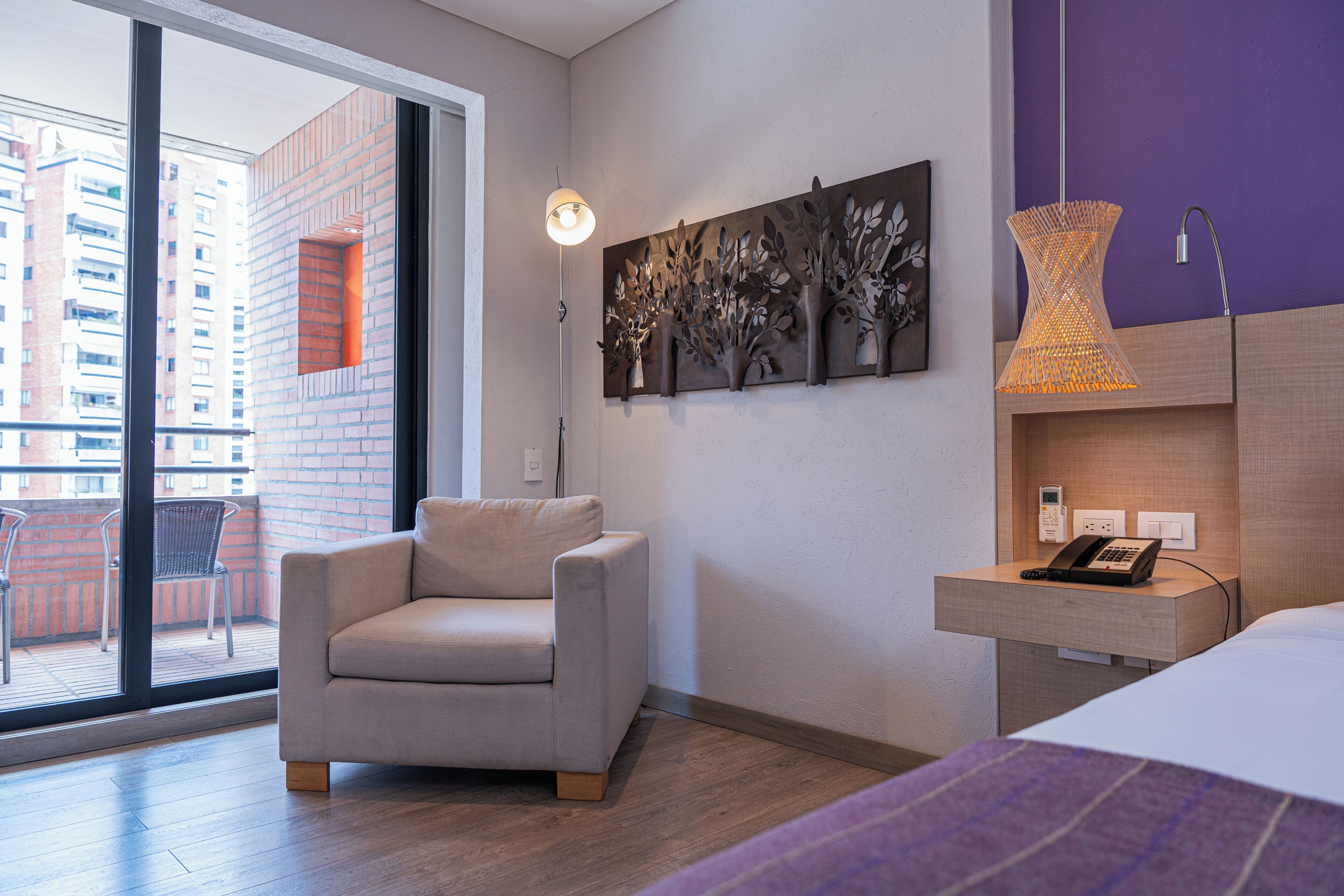 A modern hotel room with a sofa, a bed, a nightstand with a phone, a wall-mounted artwork, and a view of the outside balcony.