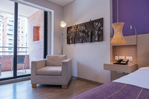 A modern hotel room with a sofa, a bed, a nightstand with a phone, a wall-mounted artwork, and a view of the outside balcony.