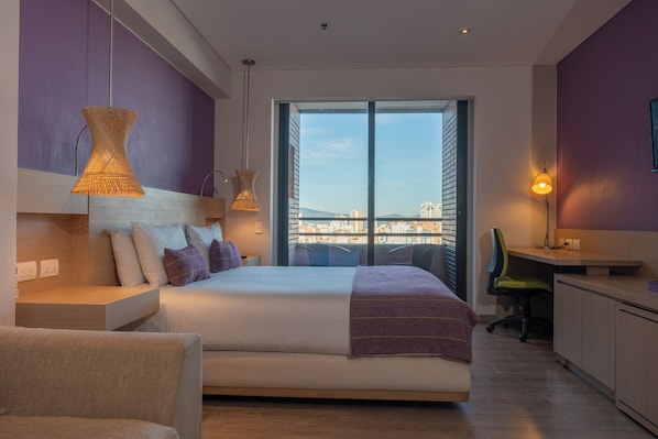 A modern hotel room with a large bed, a desk, and a view of a cityscape through a large window.