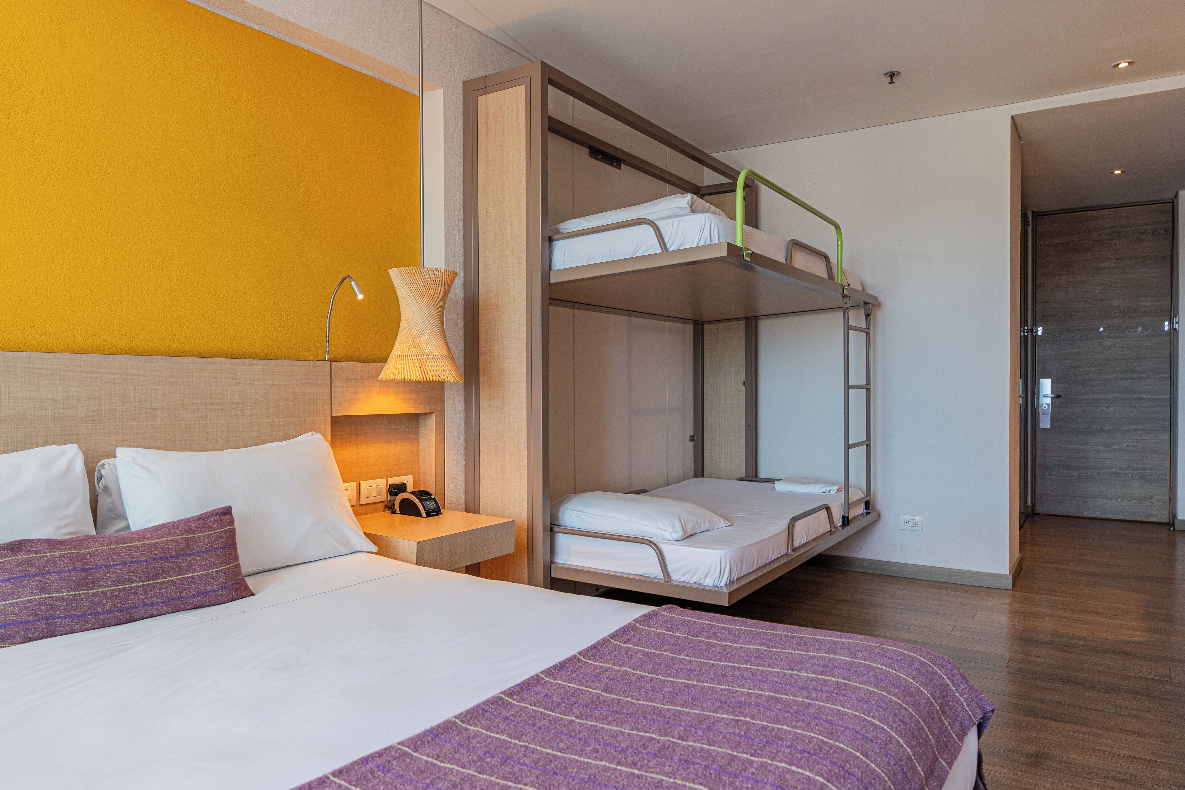 Une chambre d’hôtel avec un lit superposé, une table de chevet avec une lampe et un lit recouvert d’une couverture à rayures violettes.