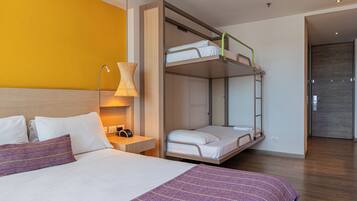 Habitación de hotel con literas, una mesita de noche con lámpara y una cama con manta a rayas moradas.