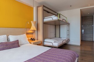 Habitación de hotel con literas, una mesita de noche con lámpara y una cama con manta a rayas moradas.