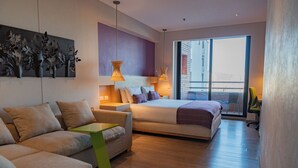 A modern hotel room with a bed, sofa, and a small table. There is a balcony with a view of the city.