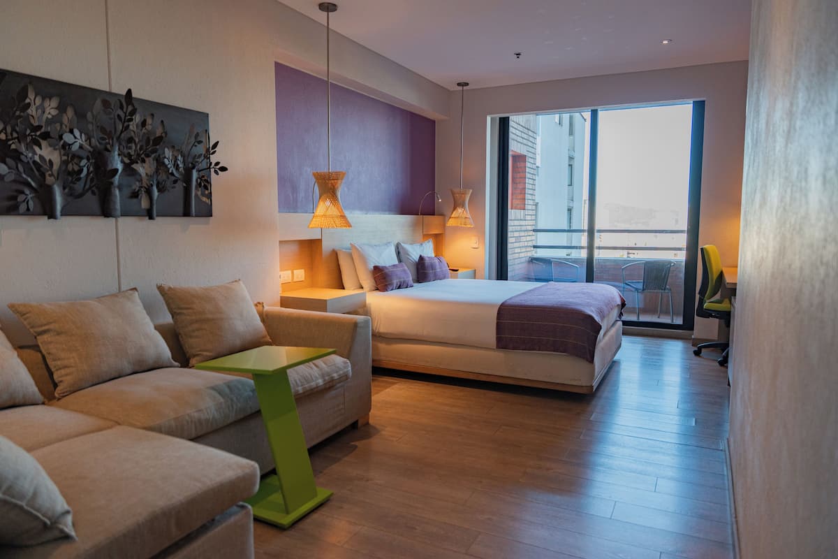 A modern hotel room with a bed, sofa, and a small table. There is a balcony with a view of the city.