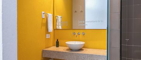 Un baño con pared amarilla, un lavabo blanco, un espejo y una zona de ducha.