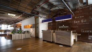 A modern hotel reception area with wooden reception desks, a high ceiling with exposed pipes, and a brick wall with the hotel's name.