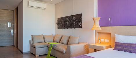 A modern hotel room with a sofa, a bed, a nightstand, and a wall-mounted artwork.