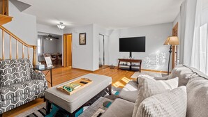 A modern living room with a staircase, a flat-screen TV, and a dining area in the background. - Cheerful 3 bedroom. Feels like home. (Chicago)