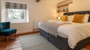 A bedroom with a bed, a chair, a window with blinds, a radiator, and a shelf with items. - 3 Settlebeck Cottages (Sedbergh)