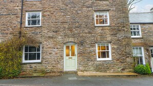 A stone building with a green door, multiple windows, and a small garden area with yellow flowers. - 3 Settlebeck Cottages (Sedbergh)