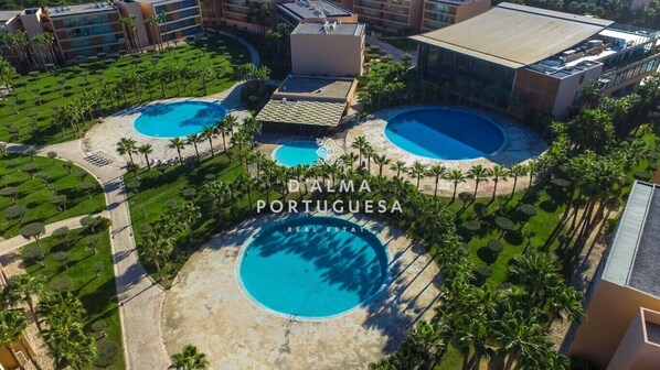 Vue aérienne d’un complexe hôtelier doté de plusieurs piscines, de palmiers et d’un bâtiment à l’architecture moderne.