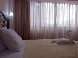 Quarto de hotel com cama, abajur, cortinas leves e toalha em formato de cisne sobre a cama.
