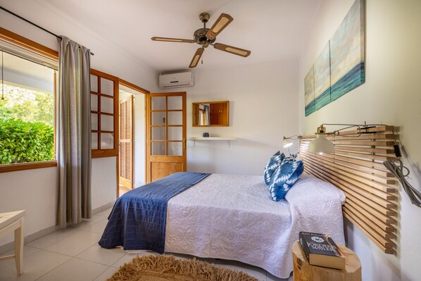 A bedroom with a bed, a ceiling fan, a window with curtains, a bedside table with a book, and a wall-mounted lamp. - Villa Vora by JS Villas (Port de Pollença)