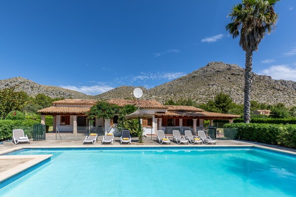 A pool with sun loungers and a house with a tiled roof.