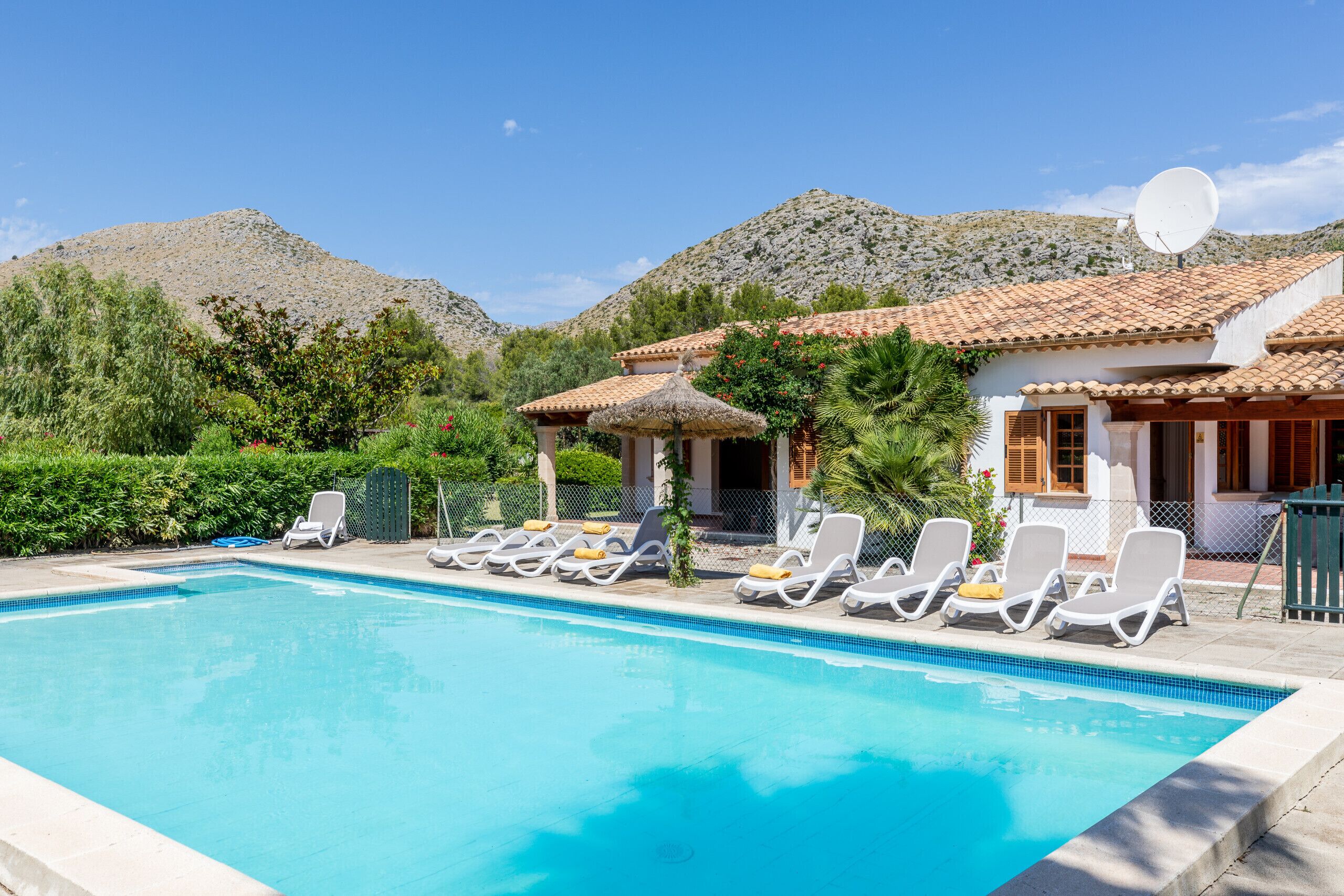 A swimming pool with lounge chairs, a building with a tiled roof, and a mountainous backdrop.