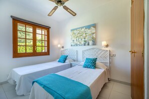 A room with two single beds, a ceiling fan, a window with a view of greenery, and a painting on the wall.