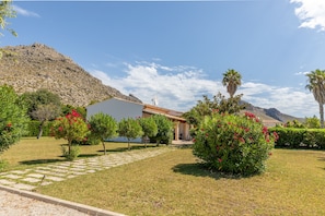 Uma casa com um caminho pavimentado que leva à entrada, cercada por vegetação bem cuidada e uma montanha ao fundo.