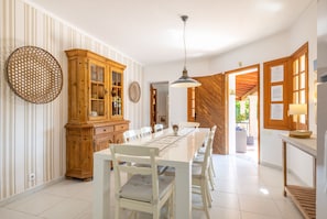 Uma área de refeições com uma mesa comprida, cadeiras brancas, um armário de madeira e um lustre.