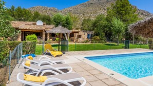 A poolside area with lounge chairs, a small building with a thatched roof, and a mountainous backdrop. - Villa Costa by JS Villas (Port de Pollença)