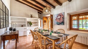 A dining area with a wooden table set for four, surrounded by wicker chairs. A white shelf unit with decorative items is visible to the left. - Villa Costa by JS Villas (Port de Pollença)