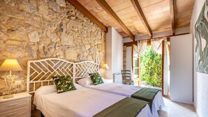 A bedroom with two beds, stone walls, wooden beams, and a view of greenery outside. - Villa Costa by JS Villas (Port de Pollença)