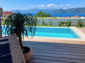 Een houten terras met een zwembad, een potplant en uitzicht op zee en bergen.