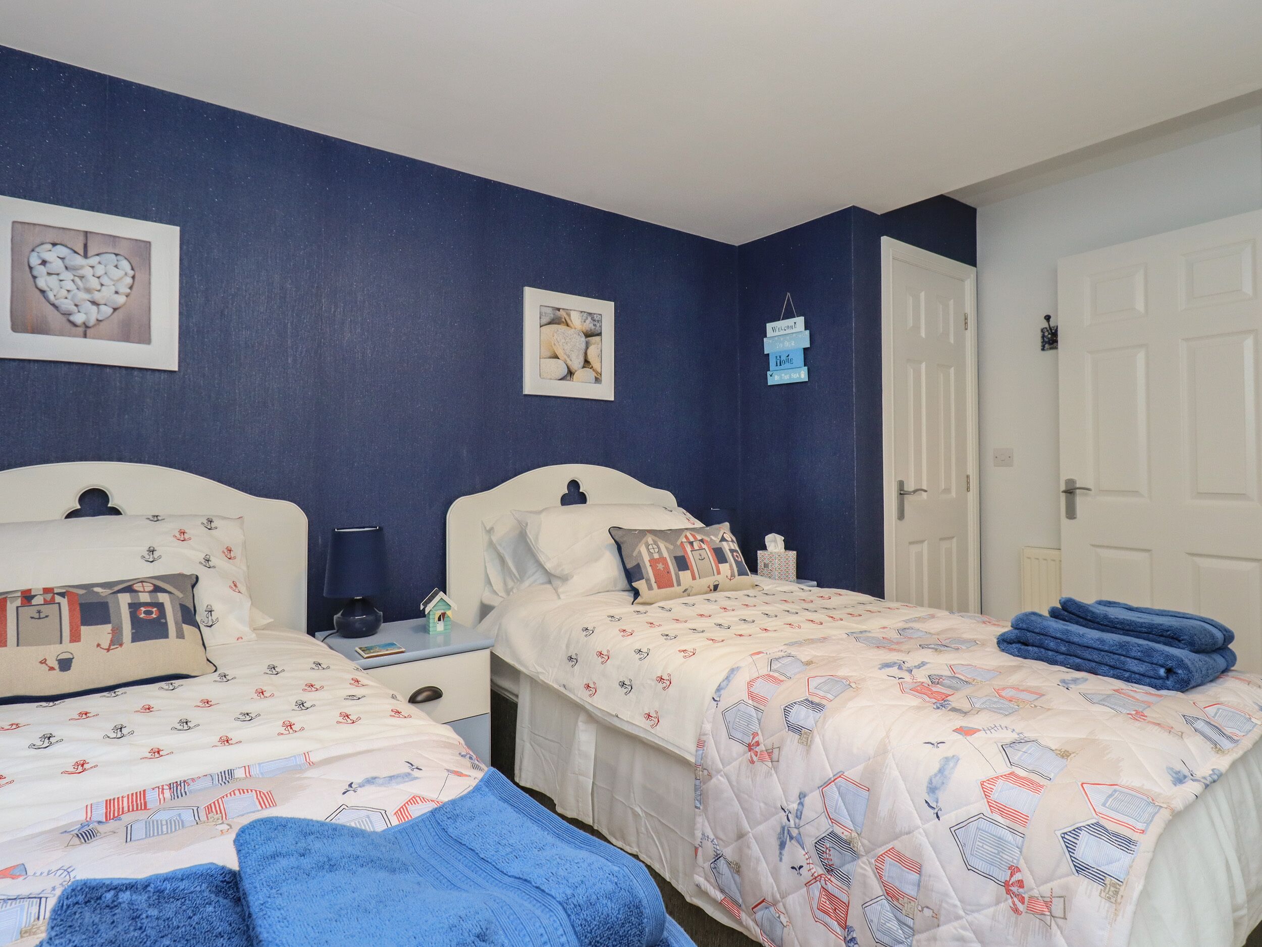 Quarto duplo com duas camas, uma mesa de cabeceira, uma luminária e uma pequena mesa. Há duas imagens emolduradas na parede e uma placa de boas-vindas.