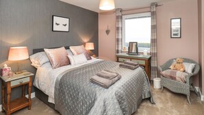 Un dormitorio con cama, dos mesitas de noche, cómoda, silla y ventana con cortinas.