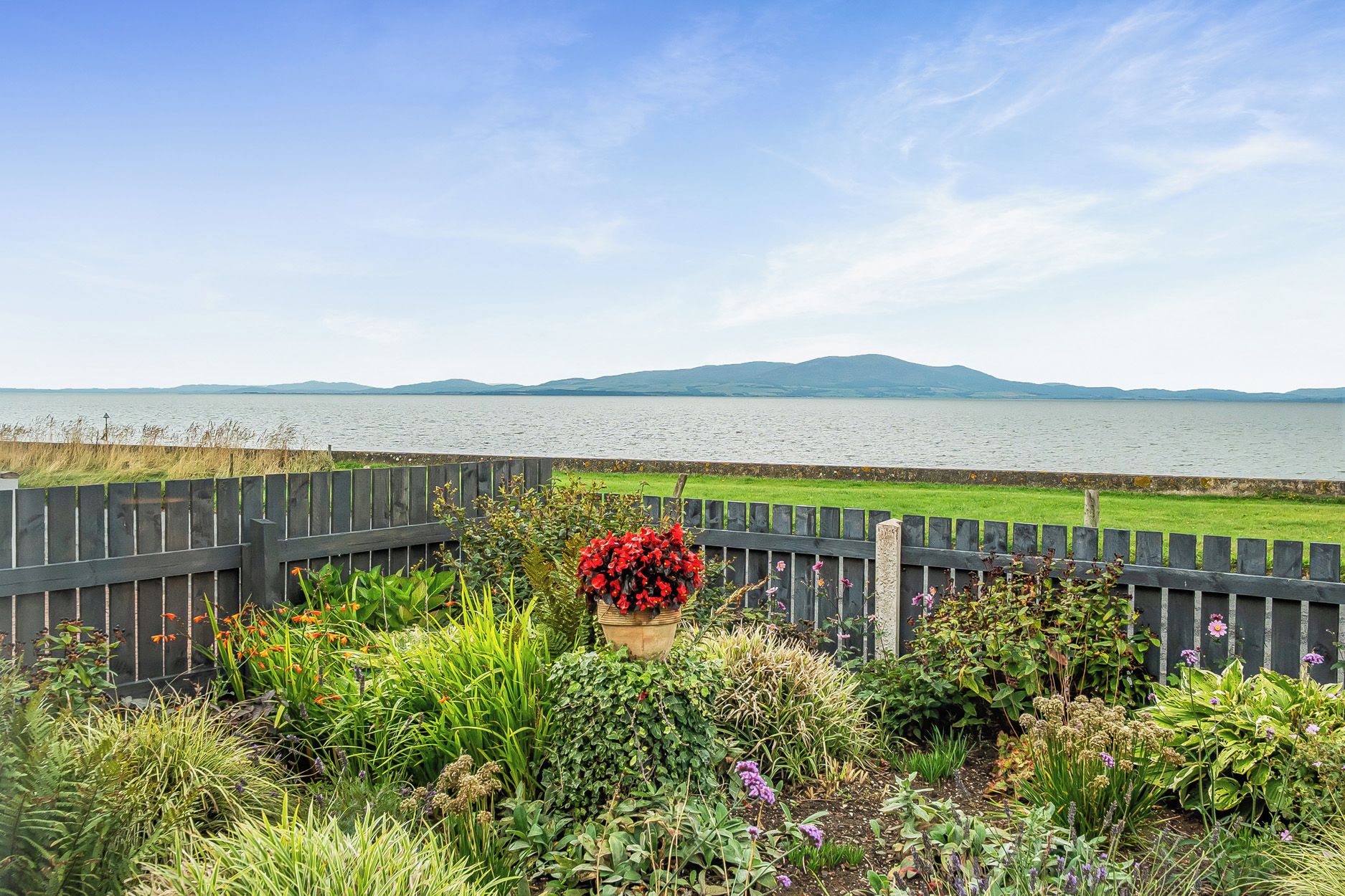 A garden with a variety of plants and flowers, a wooden fence, and a view of a lake with mountains in the distance.