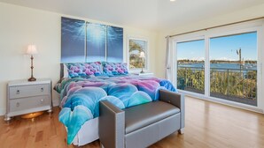 Ein Schlafzimmer mit einem großen Bett, einem Nachttisch, einer Kommode und Blick auf ein Gewässer durch große Fenster.