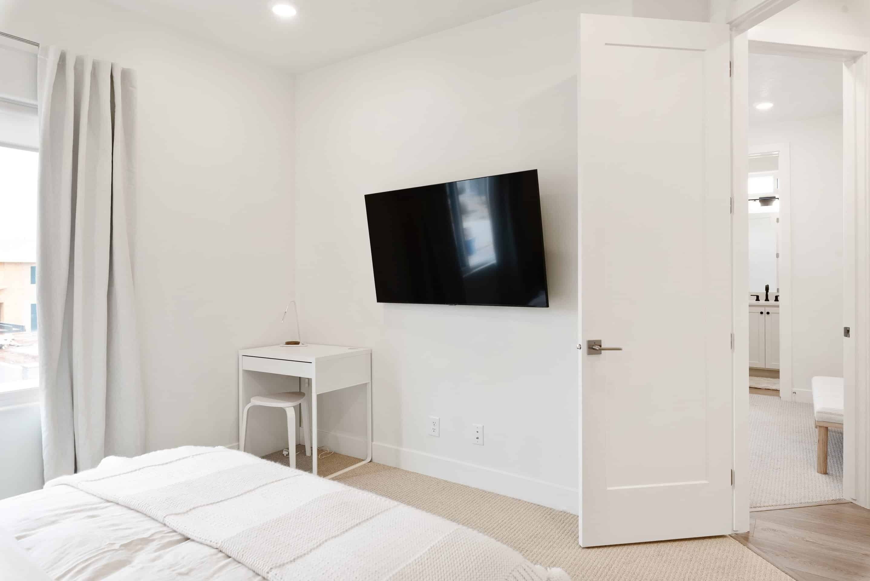 A modern bedroom with a bed, a desk, a television, and a door leading to another room.