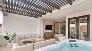 Un'area living esterna moderna con idromassaggio, divani color beige, una pergola in legno e un televisore a schermo piatto.