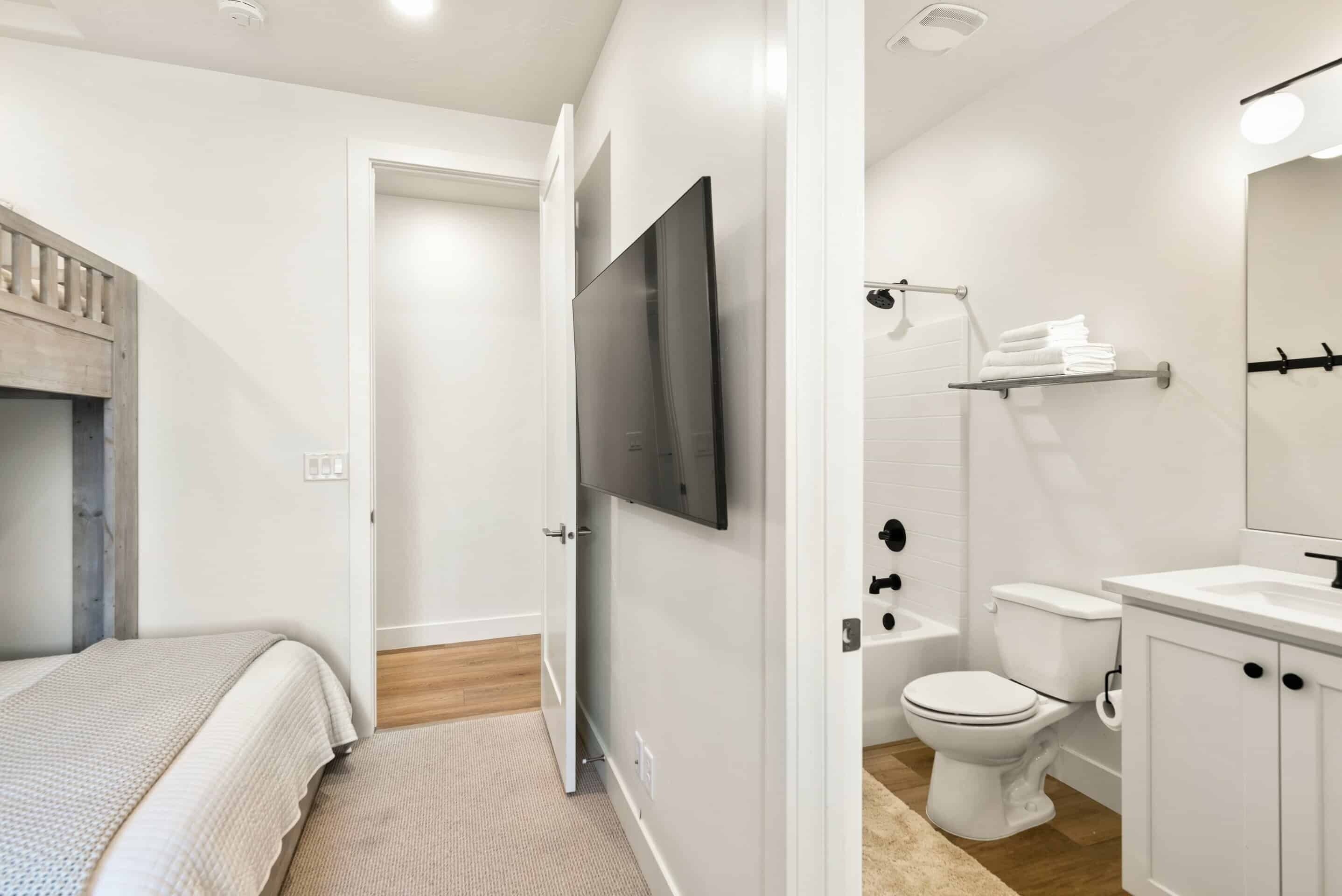 A modern bedroom with a bed, a TV mounted on the wall, and a bathroom with a toilet, sink, and towel rack.