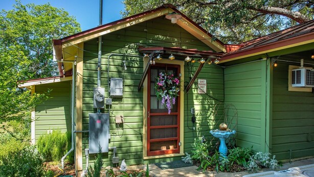 Una casa verde con puerta de madera, un pequeño porche y un jardín con un bebedero para pájaros y una fuente.