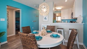 A dining area with a round table, wicker chairs, and blue tableware. - Changing Tides: Tastefully Decorated Condo with Community Pool and Easy Beach Ac (Caswell Beach)