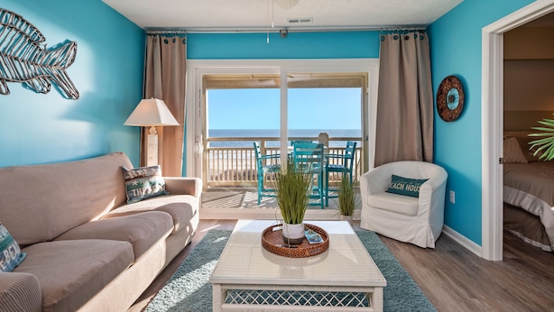 A room with a view to the sea, a ceiling fan, a sofa, a chair, and a coffee table with a plant. - Changing Tides: Tastefully Decorated Condo with Community Pool and Easy Beach Ac (Caswell Beach)
