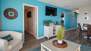 A living room with a teal wall, a white sofa, a coffee table, and a dining area with a wicker chair. - Changing Tides: Tastefully Decorated Condo with Community Pool and Easy Beach Ac (Caswell Beach)