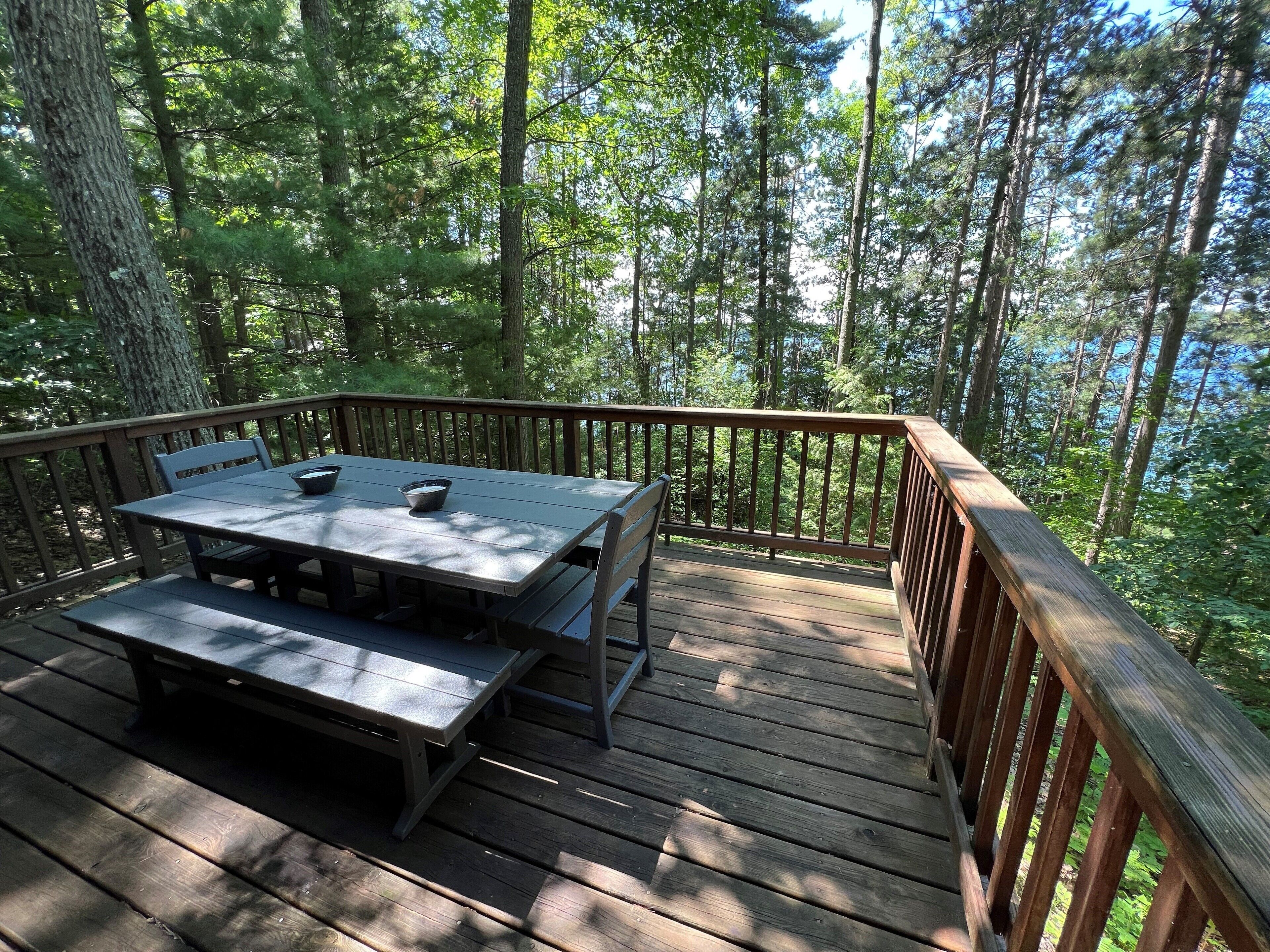 A wooden deck with a table and benches, surrounded by tall trees and a clear view of the forest.
