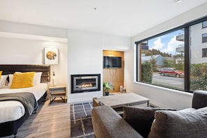 A modern hotel room with a bed, a sofa, a TV, a fireplace, and a large window with a view of the street.