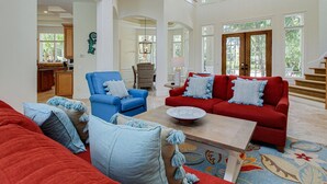 Ein geräumiges Wohnzimmer mit einem roten Sofa, einem blauen Sessel und einem Couchtisch. Im Hintergrund ist eine Küchenzeile erkennbar.