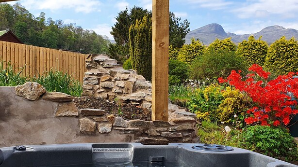 A hot tub in a garden with a wooden fence, stone wall, and colorful flowers. - Tayview (Killin)