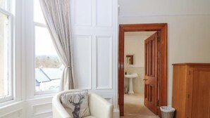 A room with a white chair, a wooden cabinet, a window with curtains, and a door leading to a bathroom. - Tayview (Killin)