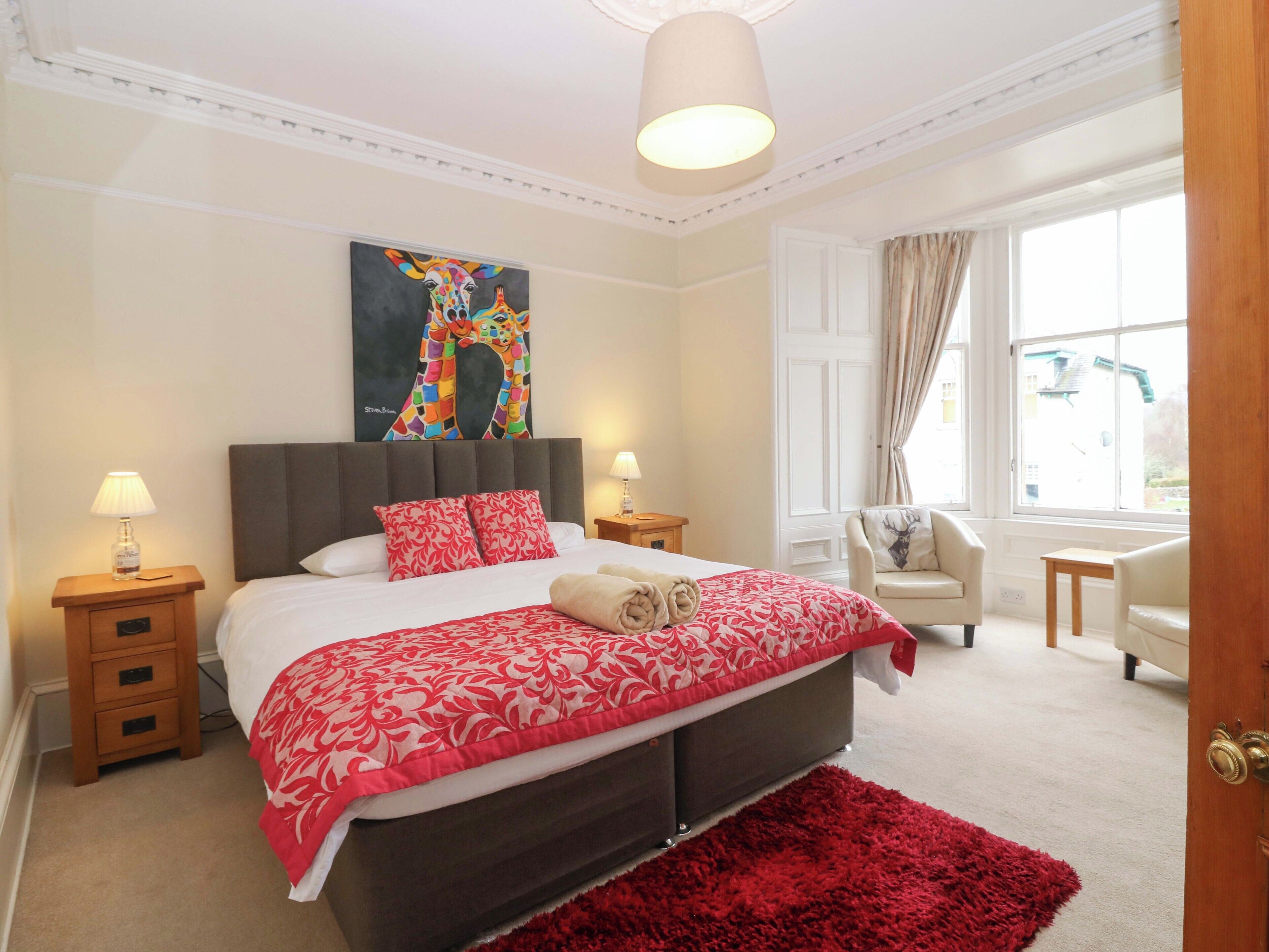 A bedroom with a large bed, two bedside tables, a chair, a window with curtains, and a painting on the wall.