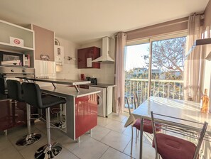 A modern kitchen with a dining area, a view of the city through large windows, and a balcony with chairs.